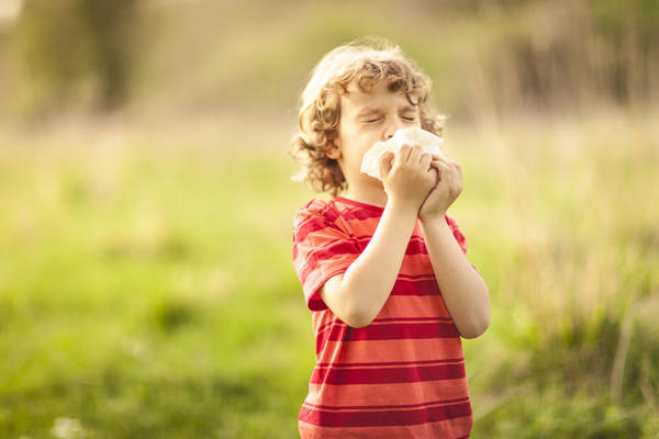 Little blond boy sneezing due to allergy related problems, on a sunny day outdoors. He is holding a handkerchief in his hands, looking away. Copy space available.