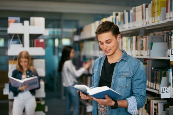Student Of University Reading Book In Library. Man With Textbook Near Bookshelves And Students On Background. High Resolution